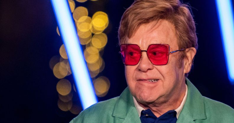 Elton John, wearing red glasses, biting his lip and looking to the right.