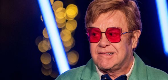 Elton John, wearing red glasses, biting his lip and looking to the right.