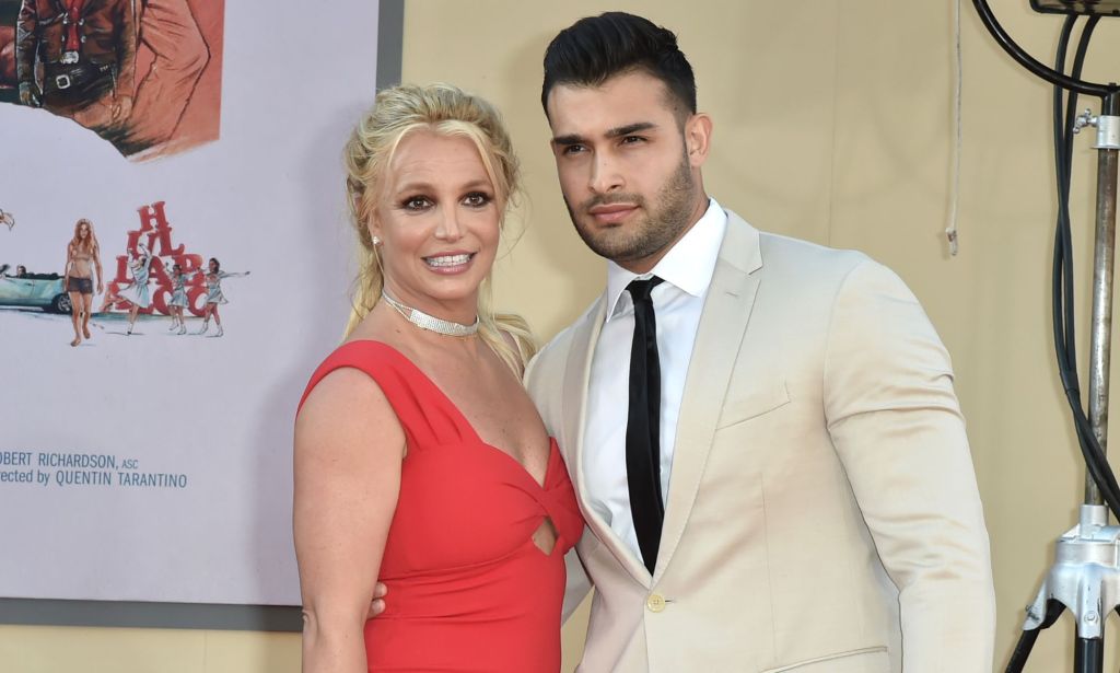 Britney Spears, wearing a red dress, and Sam Asghari, wearing a beige suit, smile for a photo outside of the Chinese Theatre in California.