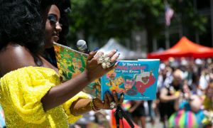 A drag queen, wearing a yellow dress, reads to a crowd from a book titled "if you're a drag queen and you know it."