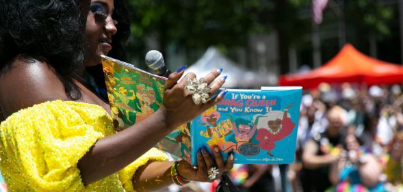 A drag queen, wearing a yellow dress, reads to a crowd from a book titled "if you're a drag queen and you know it."