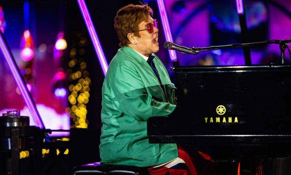 Elton John singles into a microphone while he plays on a Yamaha piano, wearing a teal suit.