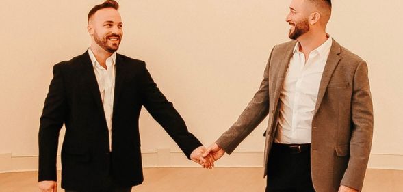 TJ House and Ryan Neitzel hold hands while looking at one another in an empty white room with wall lights above them.