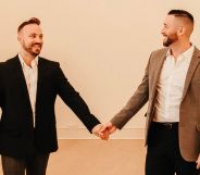 TJ House and Ryan Neitzel hold hands while looking at one another in an empty white room with wall lights above them.