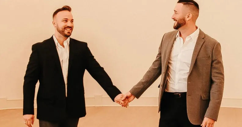 TJ House and Ryan Neitzel hold hands while looking at one another in an empty white room with wall lights above them.