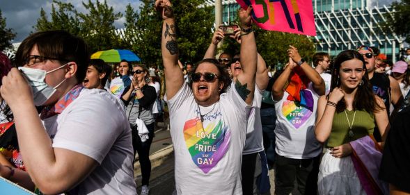 A person in a Pride shirt screams while pointing a finger in the air and holding a sign that reads "gay"