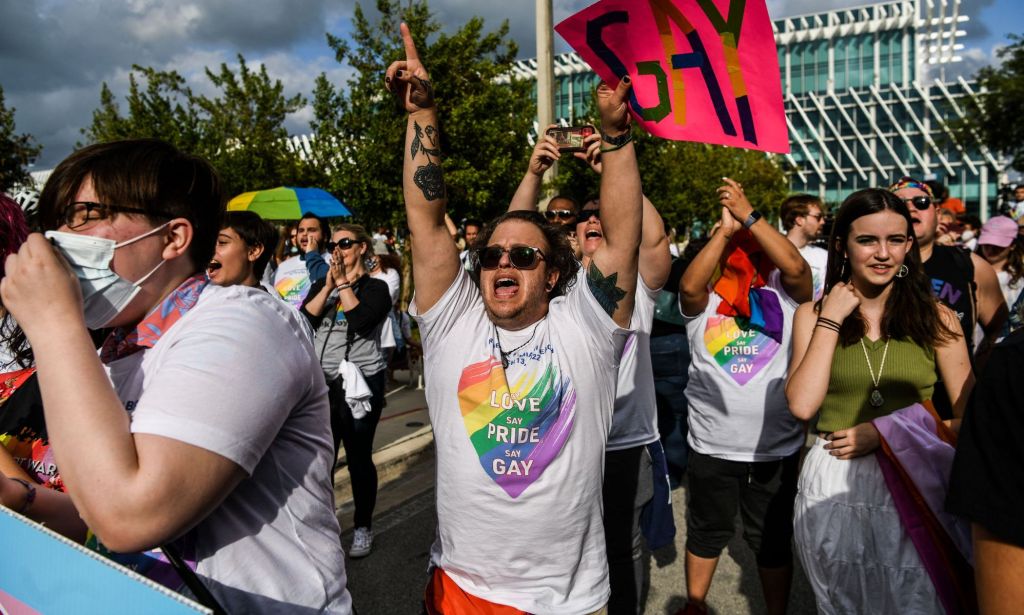 A person in a Pride shirt screams while pointing a finger in the air and holding a sign that reads "gay"