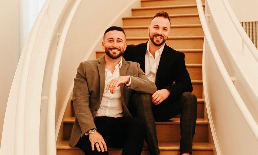 TJ House and Ryan Neitzel sit together on curved wooden stairs, leaning on one another affectionately.