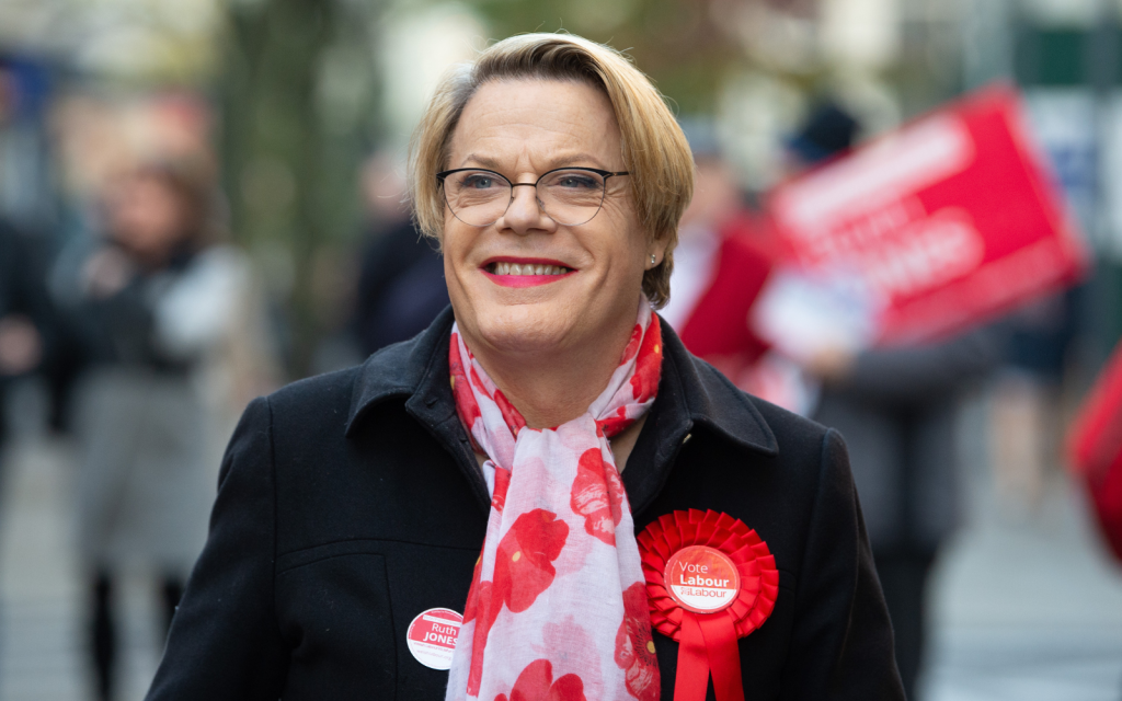 Eddie Izzard, wearing a black overcoat and a red Labour badge, walks down a local high street with fellow campaigners blurred out in the background.