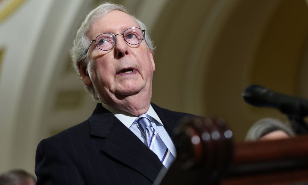 A canted shot of Mitch McConnell, in a black suit and silver tie, speaking at a stand.