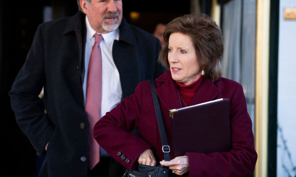 Vicky Hartzler, wearing a dark red coat, reaches into her bag while looking to the right, a tall older man stands behind her with a pink tie.