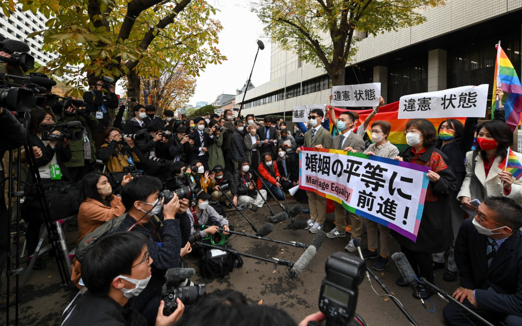 A wider view shot of photographers surrounding a pro-LGBTQ+ group waving banners in support of same-sex marriage near a Tokyo court.
