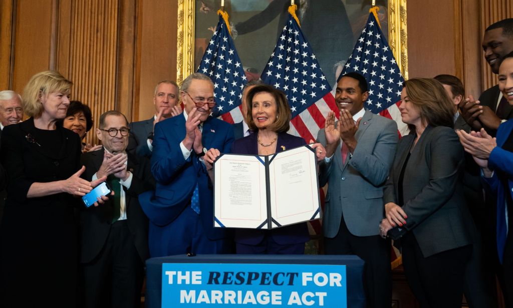 Nancy Pelosi holds up a copy of the Respect for Marriage Act, surrounded by individuals applauding.