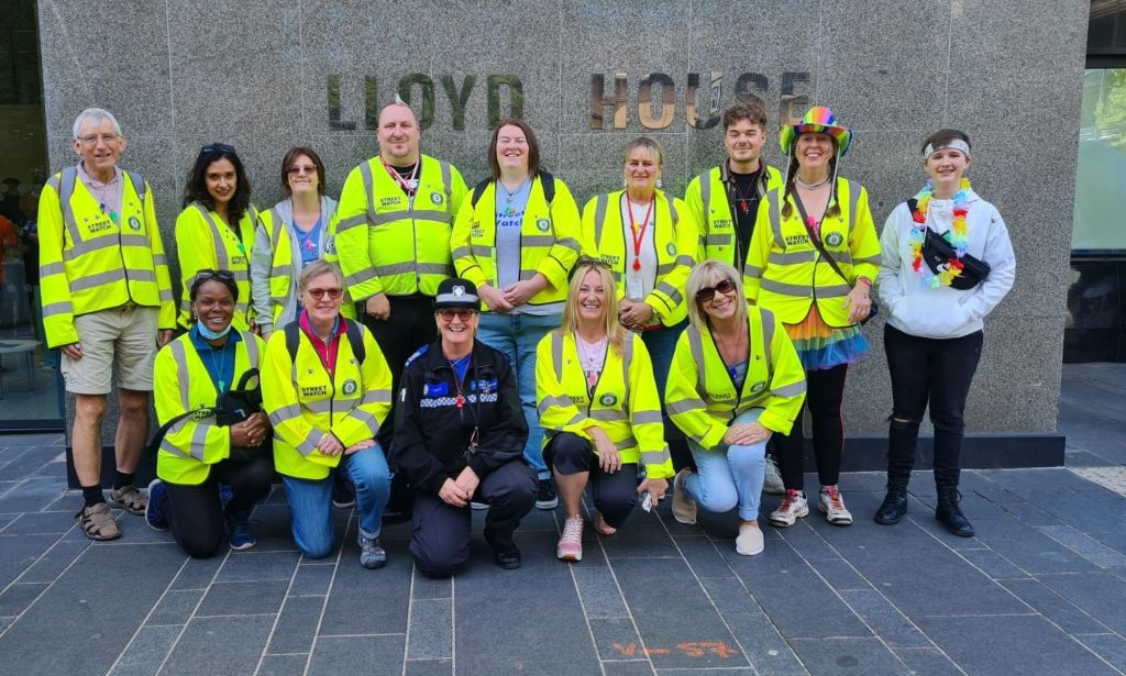 Members of the Birmingham StreetWatch crew wear yellow high-vis jackets, smiling in front of the police headquarters.