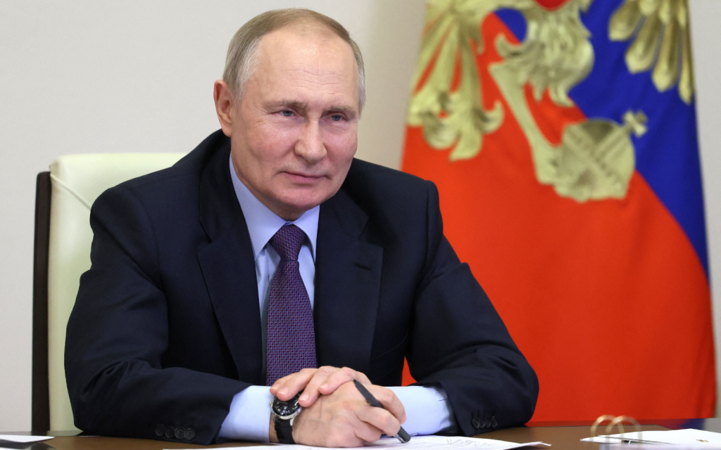 Russian president Vladimir Putin sits at a desk next to a flag with a pen in his hand.