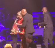 Patti LaBelle, in a red dress, is escorted off stage by two men in black suits, as the rest of the band is escorted off in the background.