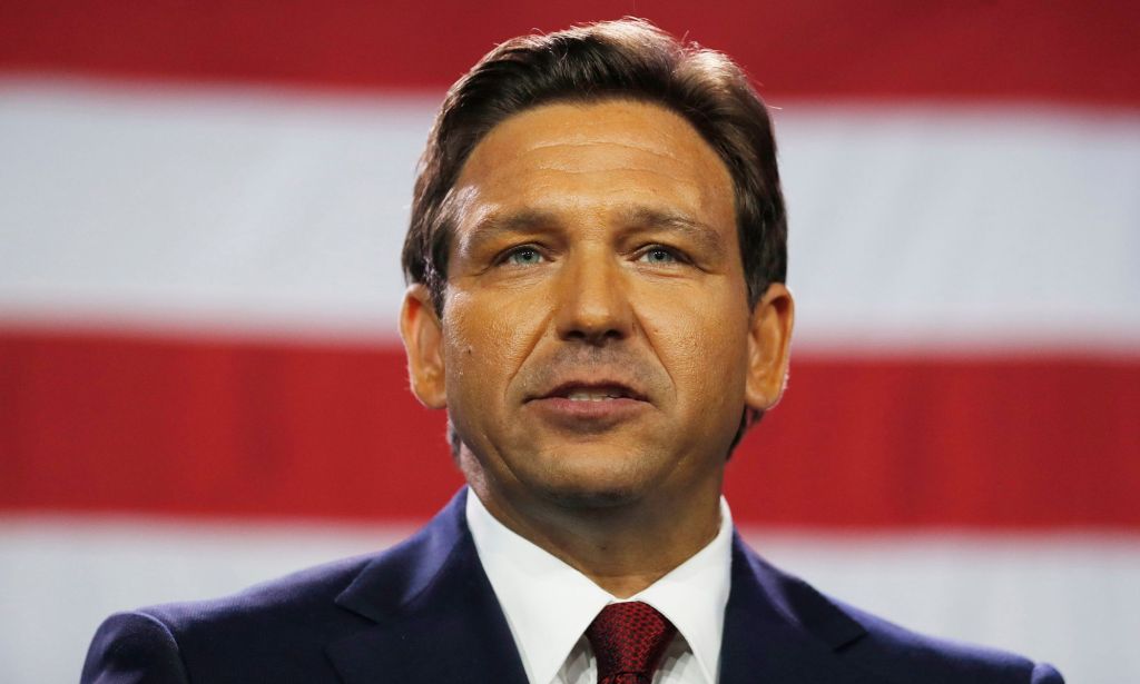 Florida governor Ron DeSantis, wearing a suit and tie, stands in front of the red and white stripes of the US flag
