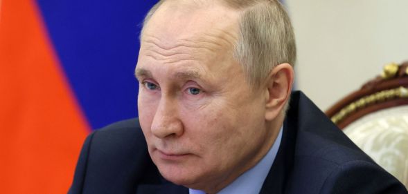 Vladimir Putin stares at someone off camera while sitting in front of a Russian flag after signing an extended LGBTQ+ 'propaganda' bill into law