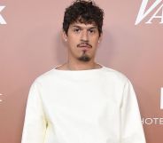 Omar Apollo wears a white top as he stares at the camera while standing in front of a rose pink background