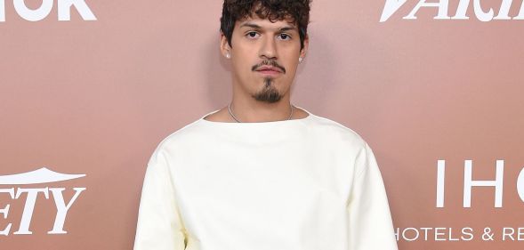 Omar Apollo wears a white top as he stares at the camera while standing in front of a rose pink background