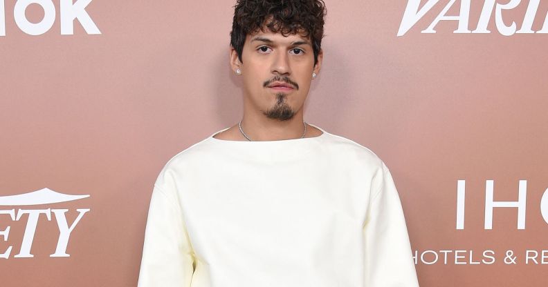 Omar Apollo wears a white top as he stares at the camera while standing in front of a rose pink background