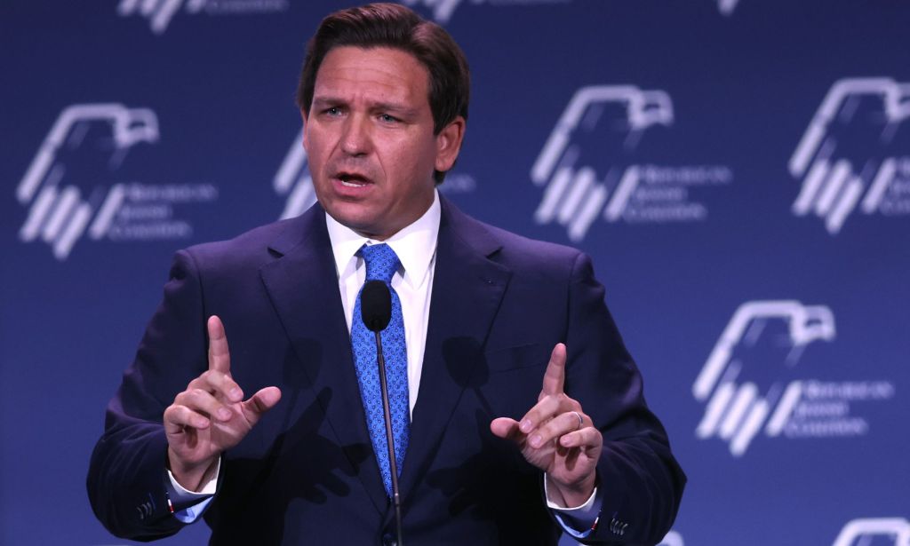 Ron DeSantis, wearing a suit and tie, gestures with both hands in the air as he speaks into a microphone at an event
