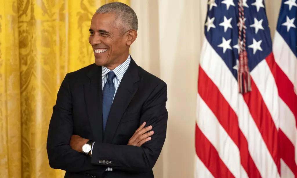Former president Barack Obama smiles as he wears a suit with his arms crossed. He is standing in front of a red, white and blue American flag