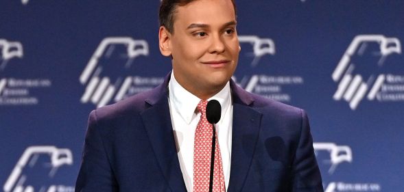 George Santos wears a suit and tie while speaking into a microphone at a conference