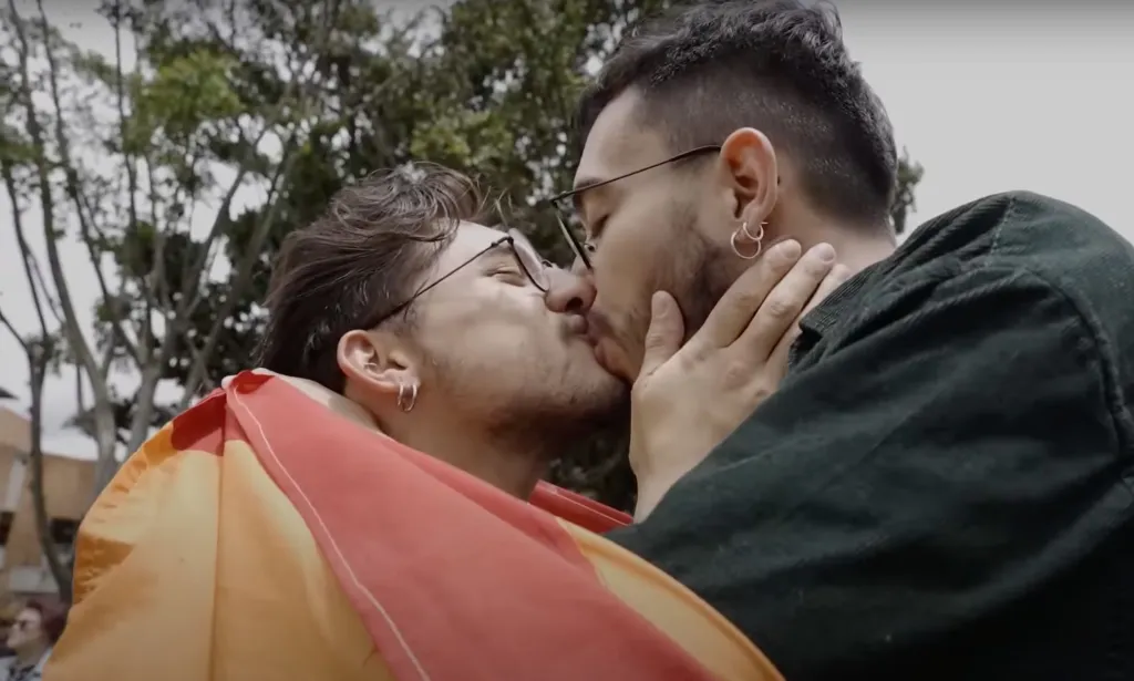 A person wearing a rainbow LGBTQ+ pride flag leans in to kiss another person