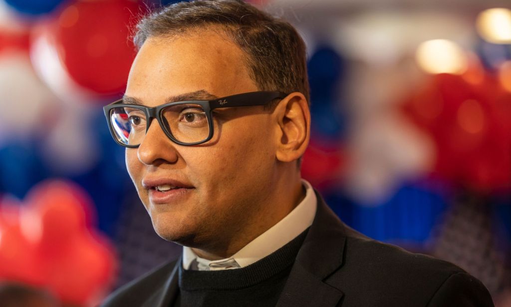 George Santos, wearing glasses and dark clothing, looks somewhere off camera during a press conference. There are red, white and blue colours in the background