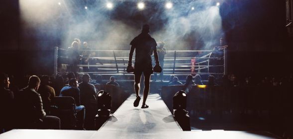 A picture shows a boxer from the back entering a boxing ring - there are crowds of people sitting either side of him