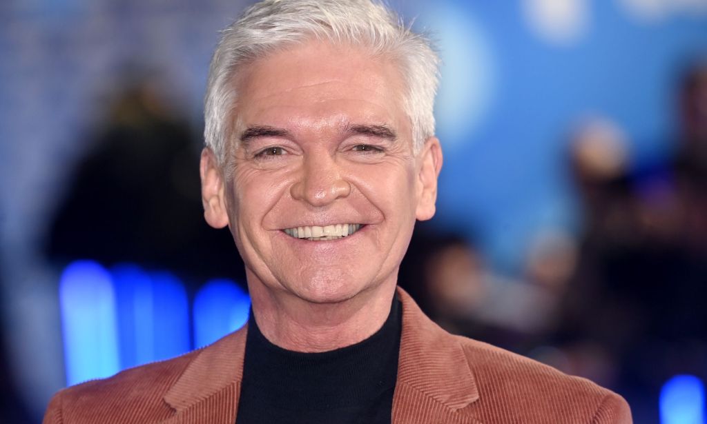 Phillip Schofield smiles at the camera while wearing a black top and brown jacket over it