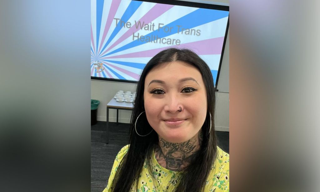 Eva Echo smiles at the camera while wearing a yellow and green patterned top with a screen in the background that reads 'The Wait For Trans Healthcare' in the colours of the trans Pride flag