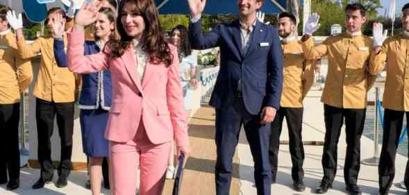 A still from HBO series The White Lotus showing characters dressed in their hotel uniforms waving - the majority wearing yellow shirts and black trousers and the two management employees wearing suits; the woman's being pink and the man's navy blue