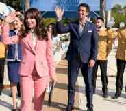 A still from HBO series The White Lotus showing characters dressed in their hotel uniforms waving - the majority wearing yellow shirts and black trousers and the two management employees wearing suits; the woman's being pink and the man's navy blue