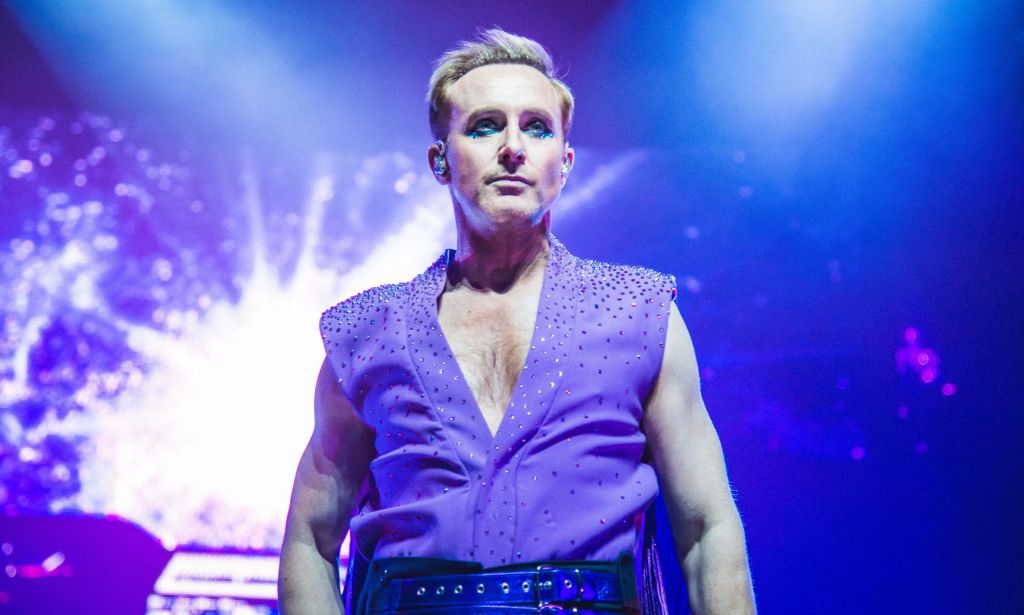 A photo of Ian "H" Watkins of Steps wearing a purple shirt with sequins as he stands on a stage with spotlights shining behind him