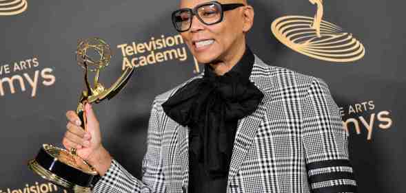 A photo of drag queen and TV personality RuPaul dressed in a grey patterned suit over a black shirt and tie with designer glasses and holding an Emmy award
