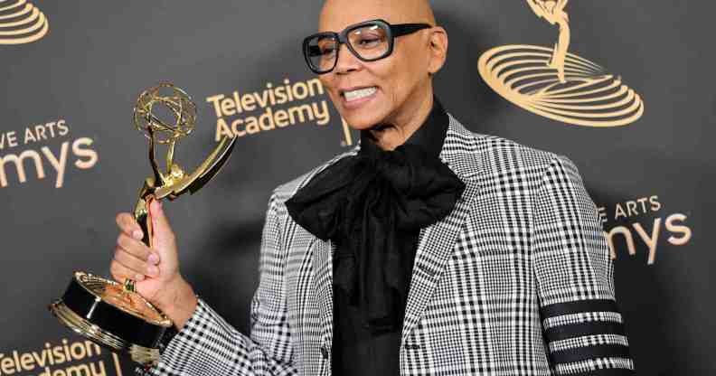 A photo of drag queen and TV personality RuPaul dressed in a grey patterned suit over a black shirt and tie with designer glasses and holding an Emmy award