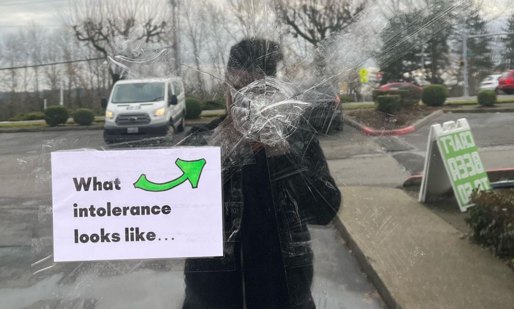A photo showing the window of a bar in Seattle that had been shot. The glass is splintered and there is a sign stuck on the window just below the point of entry that says: "What intolerance looks like"