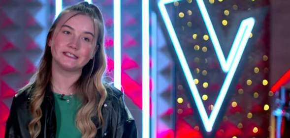A still from The Voice Kids UK shows trans contestant Darcie wearing a green top and black jacket standing on stage with a bright neon 'V' in the background