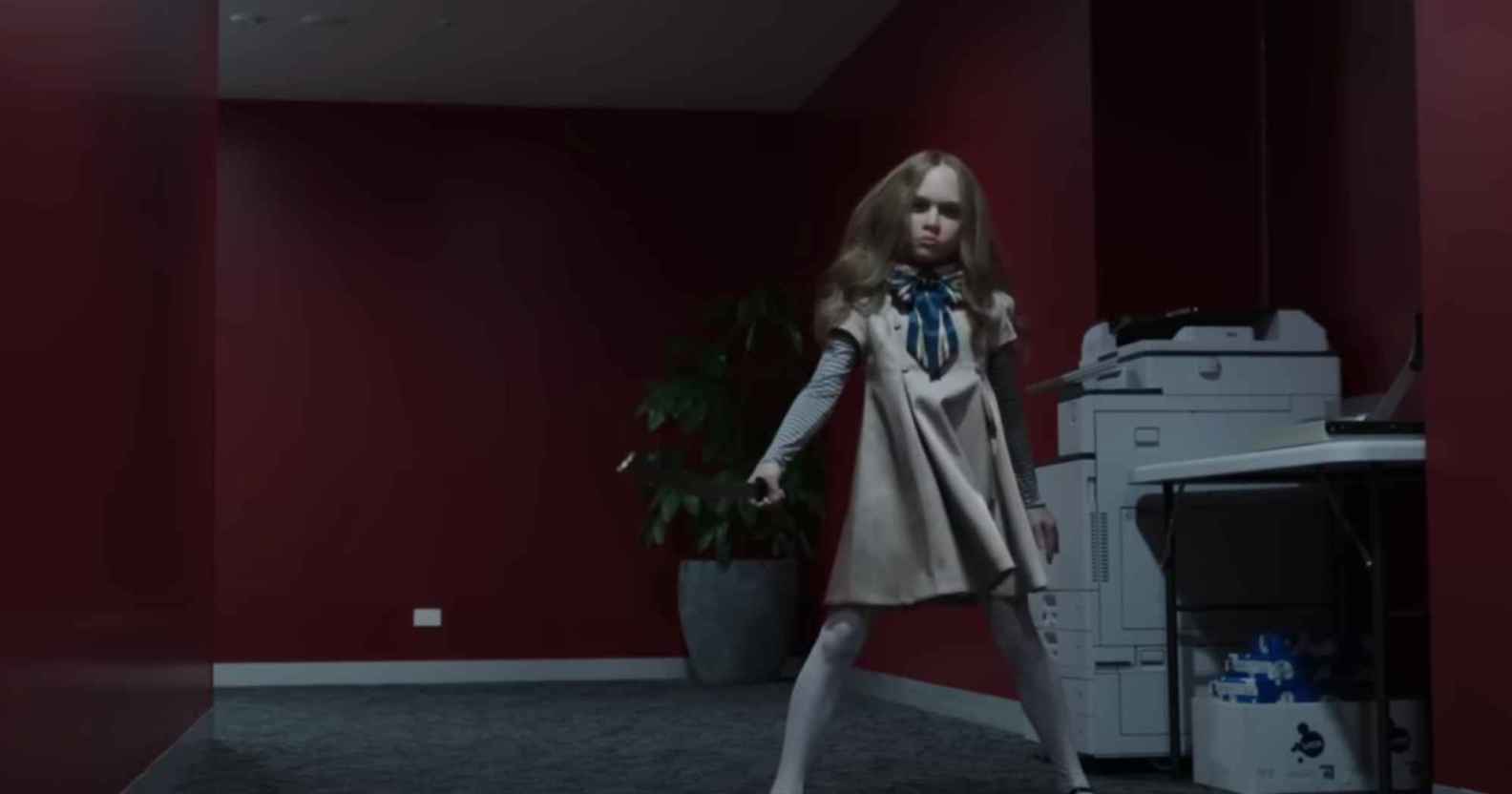 A still from horror film M3GAN showing the robot doll dancing in an office corridor that has red walls and a grey carpet. To the right of the M3GAN robot is a photocopying machine and a small table