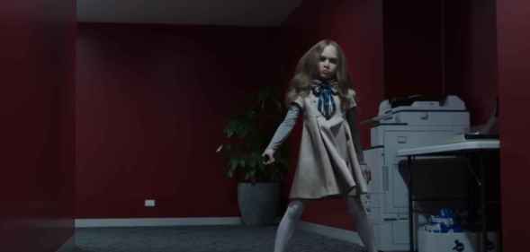 A still from horror film M3GAN showing the robot doll dancing in an office corridor that has red walls and a grey carpet. To the right of the M3GAN robot is a photocopying machine and a small table