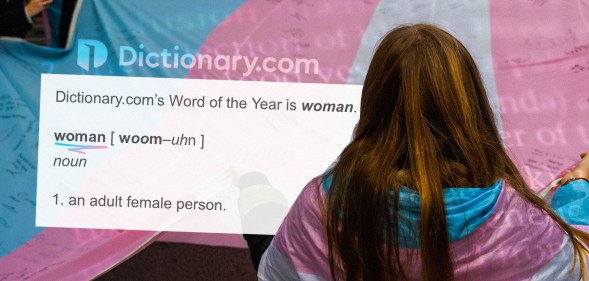 A woman stands with her back to the camera, wearing a trans flag while the dictionary definiton of the word woman is next to her.