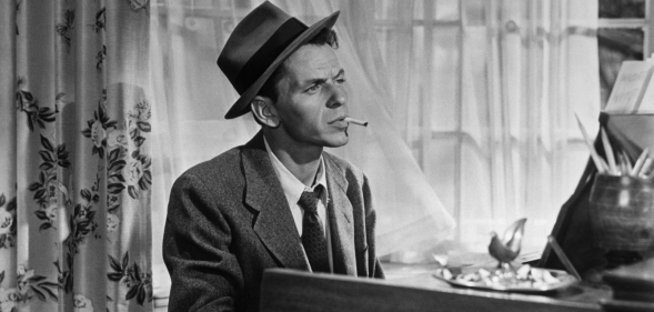 Black and White photo of Frank Sinatra sitting at a piano smoking a cigarette wearing a suit and tie.