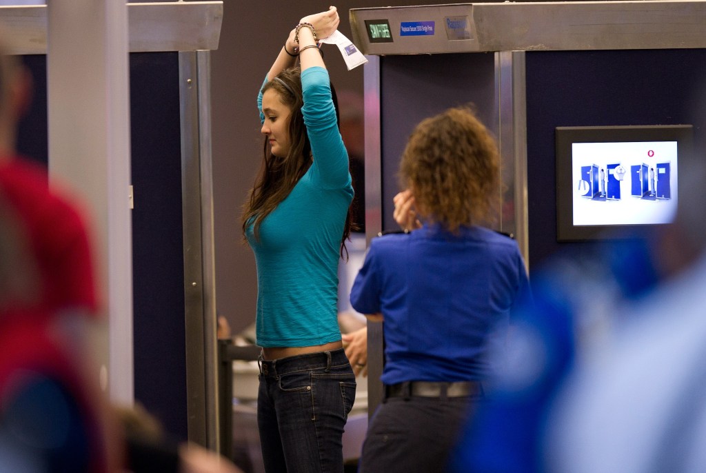 Photo of a woman wearing a turquoise-coloured top and jeans going through an airport full-body scanner with her arms raised
