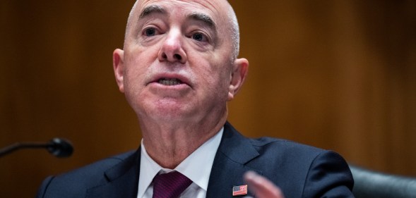 Alejandro Mayorkas wears a black suit with burgendy tie, while speaking at the Senate building in a Homeland Security meeting.