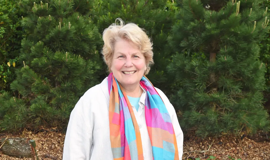 Sandi Toksvig smiles for a photo wearing a rainbow scarf, with greenery in the background
