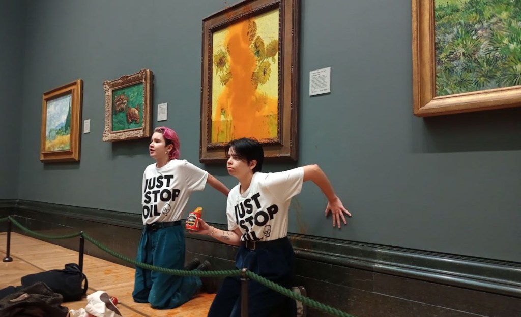 A photo of Just Stop Oil activists Anna Holland and Phoebe Plummer wearing T-shirts with the slogan "Just Stop Oil" kneel in front of Van Gogh's Sunflowers in London's National Gallery, which is covered in tomato soup