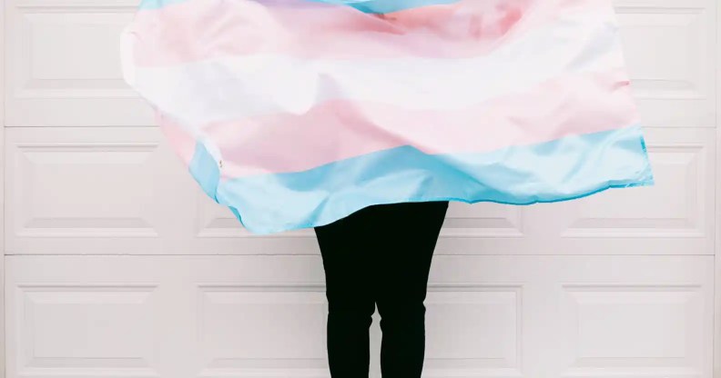 An image showing a person holding a transgender flag against a pale pink wall. The picture does not show the person's face