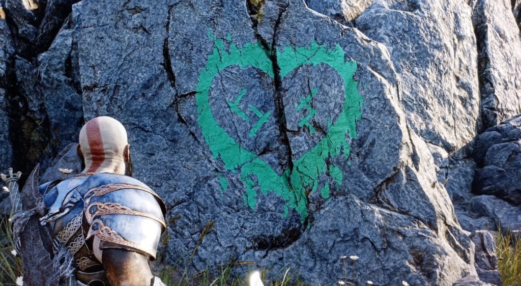 A screenshot of the game God of War Ragnarok, where the player looks at a green heart symbol painted on the side of a rock, containing Nordic symbols that translate to "SH" and "JS"